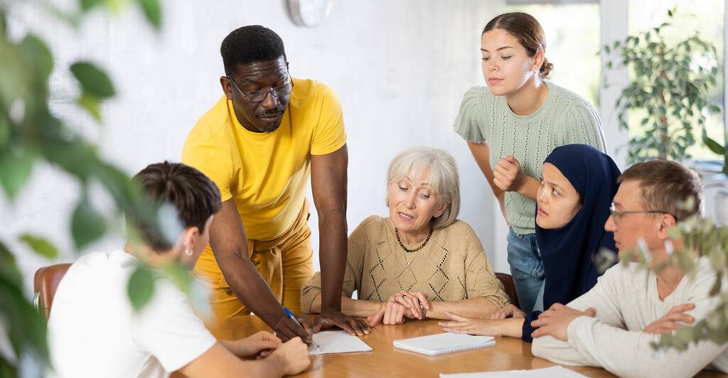 Lerngruppe Erwachsene Gruppe von Menschen verschiedener Herkunft und unterschiedlichen Alters, die um einen Tisch sitzen oder stehen und sich Notizen machen.